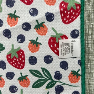 Puede incluir: Una toalla de cocina blanca con un borde verde presenta un patr&oacute;n repetido de fresas rojas y naranjas, ar&aacute;ndanos y hojas verdes. El texto "skogsbarn" est&aacute; impreso en la toalla junto con las instrucciones de cuidado y la informaci&oacute;n sobre la composici&oacute;n del material: 80% poli&eacute;ster, 20% poliamida. La toalla est&aacute; dise&ntilde;ada en Alaska y fabricada en China.