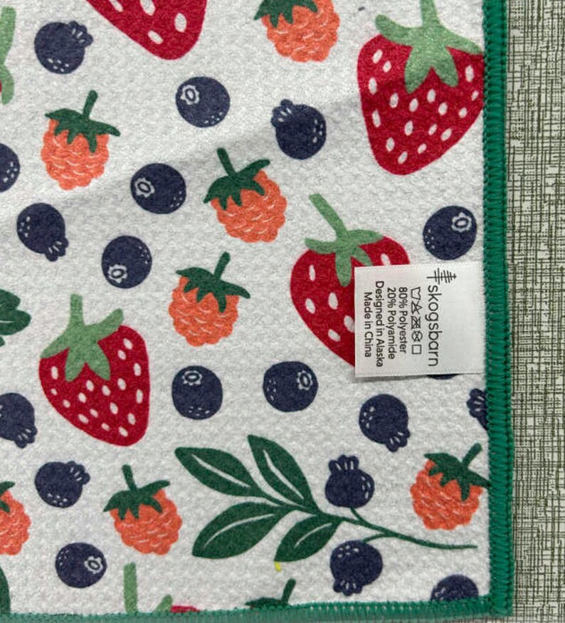 Puede incluir: Una toalla de cocina blanca con un borde verde presenta un patr&oacute;n repetido de fresas rojas y naranjas, ar&aacute;ndanos y hojas verdes. El texto "skogsbarn" est&aacute; impreso en la toalla junto con las instrucciones de cuidado y la informaci&oacute;n sobre la composici&oacute;n del material: 80% poli&eacute;ster, 20% poliamida. La toalla est&aacute; dise&ntilde;ada en Alaska y fabricada en China.