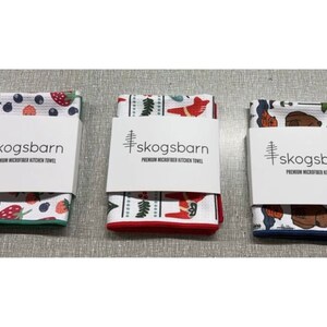 Puede incluir: Tres toallas de cocina con diferentes dise&ntilde;os. La primera toalla tiene un fondo blanco con bayas rojas y azules, hojas verdes y fresas. La segunda toalla tiene un fondo blanco con un dise&ntilde;o escandinavo rojo y blanco. La tercera toalla tiene un fondo blanco con un dise&ntilde;o azul y marr&oacute;n de un oso y &aacute;rboles. Todas las toallas tienen el texto "skogsbarn PREMIUM MICROFIBER KITCHEN TOWEL" impreso en ellas.