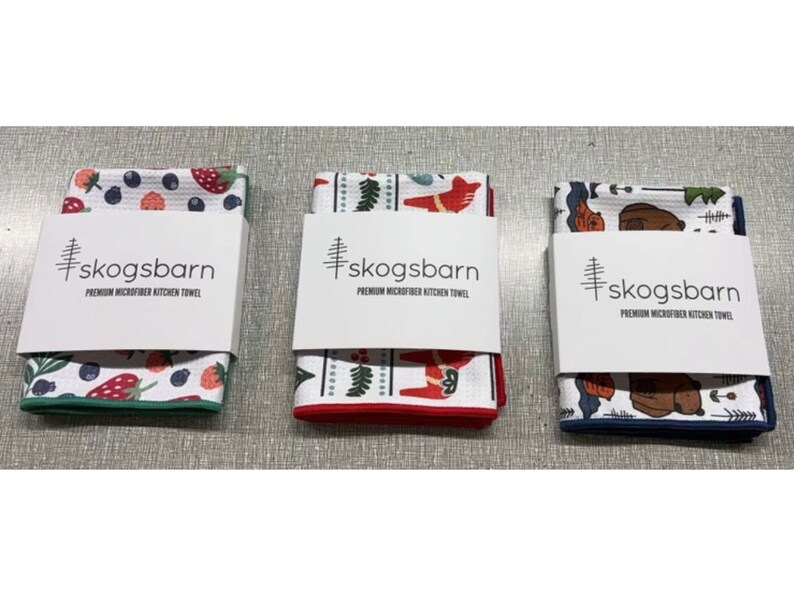 Puede incluir: Tres toallas de cocina con diferentes dise&ntilde;os. La primera toalla tiene un fondo blanco con bayas rojas y azules, hojas verdes y fresas. La segunda toalla tiene un fondo blanco con un dise&ntilde;o escandinavo rojo y blanco. La tercera toalla tiene un fondo blanco con un dise&ntilde;o azul y marr&oacute;n de un oso y &aacute;rboles. Todas las toallas tienen el texto "skogsbarn PREMIUM MICROFIBER KITCHEN TOWEL" impreso en ellas.