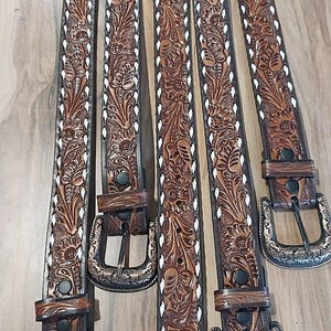 Puede incluir: Cinco cinturones de cuero marrón con intrincados diseños florales y costuras blancas en los bordes. Cada cinturón tiene una hebilla metálica decorativa. Los cinturones están dispuestos verticalmente.