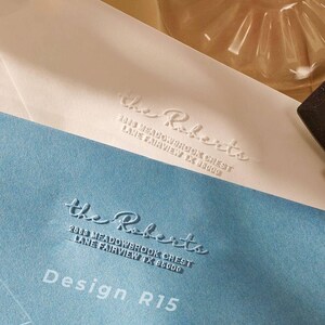 Peut inclure: Deux enveloppes, une bleu clair et une blanc cassé, avec le texte en relief "the Roberts" et les coordonnées. Un tampon noir et un verre décoratif avec un liquide rose sont également visibles. Le design est étiqueté "Design R15".
