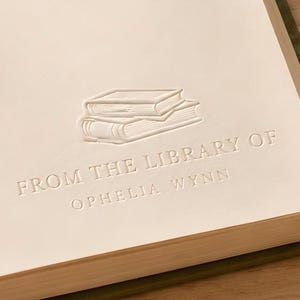 May include: A cream-coloured page featuring an embossed graphic of two stacked books above the text FROM THE LIBRARY OF OPHELIA WYNN. A gold and white fountain pen rests near the edge of the page, which is set against a light wooden surface.