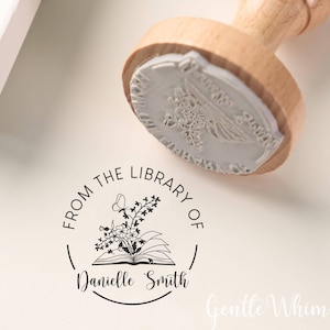 Può includere: Un timbro con manico in legno con un design floreale e di libro. Il timbro recita "From the Library of Danielle Smith" in un layout circolare. Il timbro è su una superficie color crema.