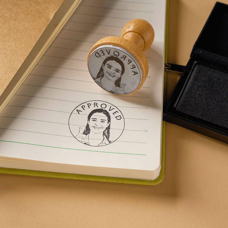 May include: A wooden stamp with a black and white image of a person giving a thumbs up. The stamp is on a lined notebook page and says "APPROVED" in a circle around the image.