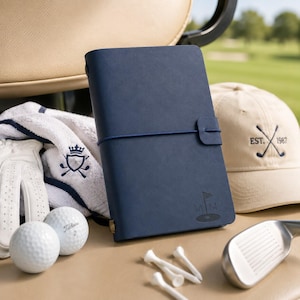 Könnte beinhalten: Ein marineblaues, ledergebundenes Notizbuch mit elastischem Verschluss, das ein geprägtes Golf-Flaggen-Logo und die Buchstaben M M zeigt. Das Notizbuch ist umgeben von einem weißen Golfhandschuh, zwei Golfbällen, einem weißen Handtuch, einer beigen Kappe und Golf-Tees.