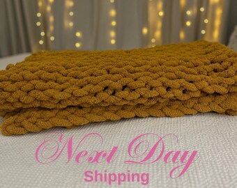 May include: A mustard yellow chunky knit throw blanket. The blanket is folded in half and placed on a white surface. The background is out of focus and shows a white curtain with string lights.