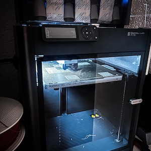 Puede incluir: Impresora 3D Bambu Lab P1S negra con puerta frontal transparente. Cuenta con un panel de control con pantalla y botones. La sección superior alberga paquetes desecantes. La placa de construcción mide 256x256x256 mm.