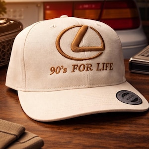 May include: A beige baseball cap with the Lexus logo embroidered in brown, and the text "90's FOR LIFE". The cap has a curved brim and a black tag on the side. The cap is sitting on a wooden surface.