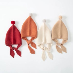 May include: Four knitted baby hats in red, orange, cream, and tan. Each hat has a pointed top with a pom-pom and ear flaps that tie under the chin. The hats are displayed on a white background.