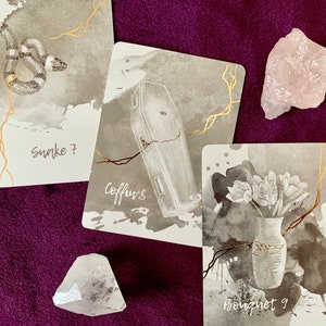 May include: Three tarot cards with black and white illustrations on a purple background. The cards are labeled "Snake 7", "Coffin 8", and "Bouqnet 9".