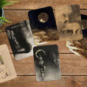 Puede incluir: Un conjunto de cartas de tarot con ilustraciones y texto, incluyendo "BLACK SMOKE", "CHIRON" y "CONSTRUCTION". Las cartas presentan im&aacute;genes oscuras con detalles dorados, sobre una superficie de madera con una peque&ntilde;a planta en maceta.