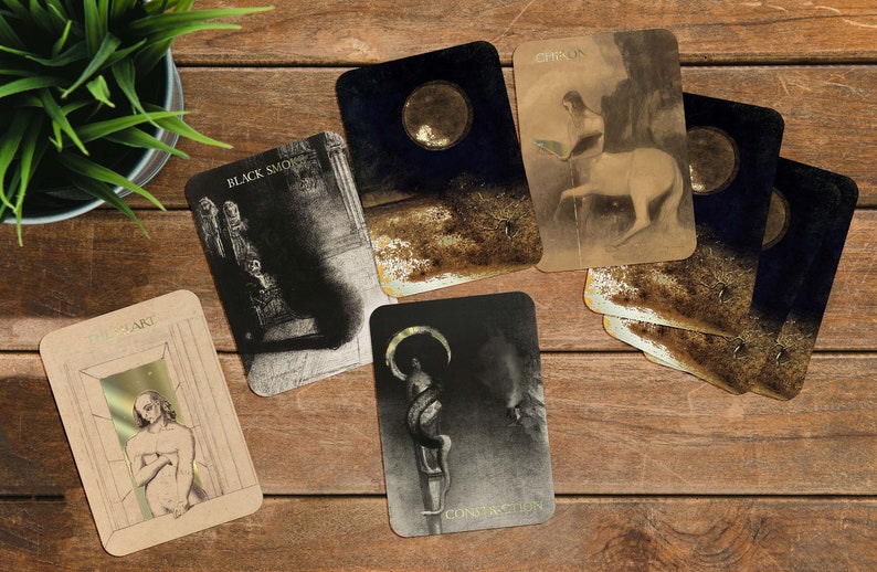 Puede incluir: Un conjunto de cartas de tarot con ilustraciones y texto, incluyendo "BLACK SMOKE", "CHIRON" y "CONSTRUCTION". Las cartas presentan im&aacute;genes oscuras con detalles dorados, sobre una superficie de madera con una peque&ntilde;a planta en maceta.
