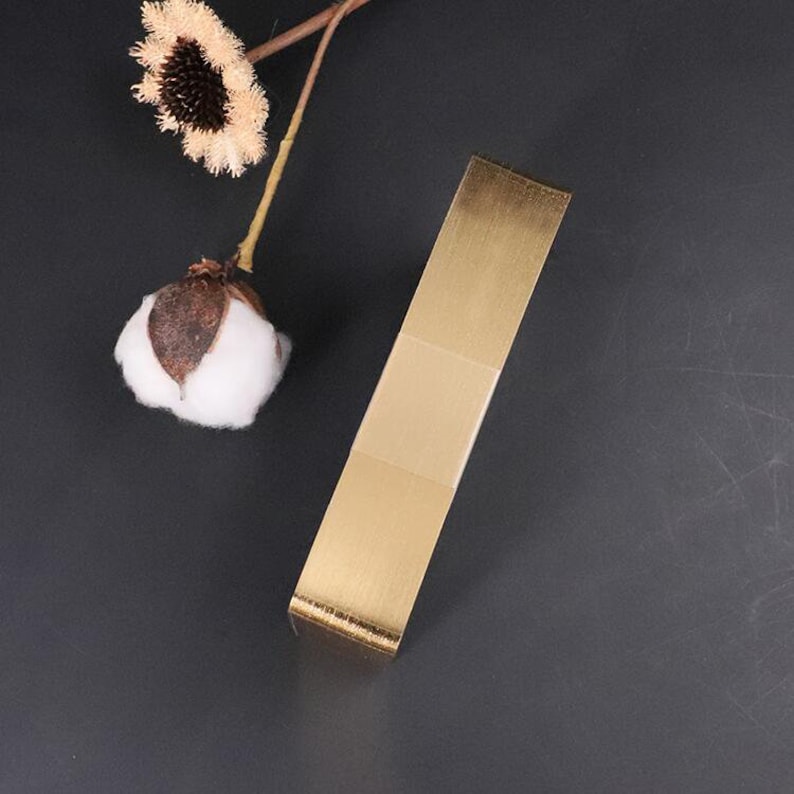 May include: A gold-colored metal door handle with a brushed finish. The handle is rectangular in shape and has a slight curve at the top.