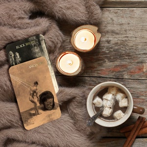 Puede incluir: Una escena acogedora con una taza de chocolate caliente con malvaviscos, dos velas encendidas en soportes de madera, cartas del tarot con el texto "BLACK SMOKE" y "ANCESTORS", y palitos de canela, todo sobre una suave manta de piel marr&oacute;n y una superficie de madera.