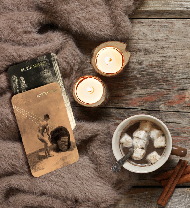 Puede incluir: Una escena acogedora con una taza de chocolate caliente con malvaviscos, dos velas encendidas en soportes de madera, cartas del tarot con el texto "BLACK SMOKE" y "ANCESTORS", y palitos de canela, todo sobre una suave manta de piel marr&oacute;n y una superficie de madera.