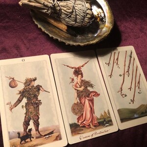 May include: Three tarot cards on a burgundy surface. The cards are from the  'Wild Unknown' tarot deck. The cards are 'The Fool', 'Queen of Pentacles', and 'Eight of Wands'.