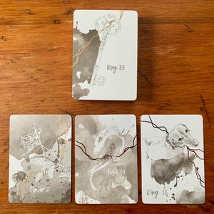 May include: Four cards with black and white illustrations and gold accents. The cards are on a wooden surface. The cards are labeled "Clover 2", "Fish 34", "Dog 18", and "Key 33".