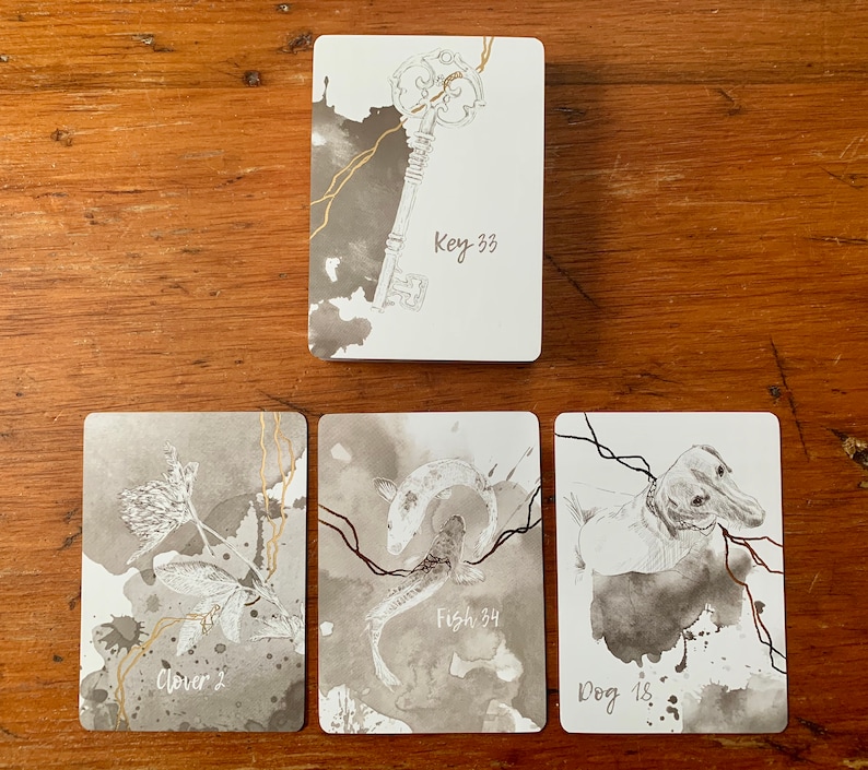 May include: Four cards with black and white illustrations and gold accents. The cards are on a wooden surface. The cards are labeled "Clover 2", "Fish 34", "Dog 18", and "Key 33".
