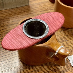 Puede incluir: Una taza de té de madera con asa y un colador de té rojo de forma ovalada. El colador tiene una malla de acero inoxidable y se asienta sobre la taza. La taza y el colador son de madera y se exhiben sobre una superficie de madera.