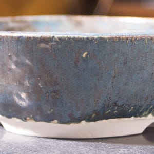 May include: A handmade ceramic bowl with a blue and brown glaze. The bowl has a wide, shallow shape and rests on a white base. The interior has a lighter blue hue, and the rim has a darker brown colour.
