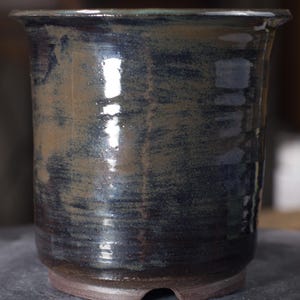 May include: A handcrafted ceramic pot with a dark blue and brown glaze. The pot has a wide mouth and a slightly flared rim. The exterior has a glossy finish with visible brushstrokes, showcasing the artisan's technique. The base is a contrasting reddish-brown.