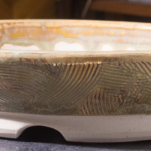 Peut inclure: Un bol en céramique fait à la main avec un motif texturé et sculpté. Le bol présente une base blanche et un bord avec un mélange de teintes vertes, brunes et jaunes. L'extérieur du bol a une texture unique et à motifs.