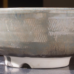 May include: A handmade ceramic bowl with a wide, shallow design. The bowl has a textured surface with diagonal lines and a grey, blue, and brown colour scheme. It rests on a white, rounded base.