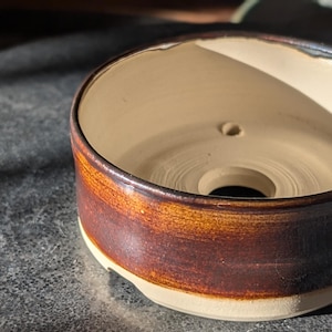 Peut inclure: Un pot en céramique brun et blanc avec un trou de drainage au fond. Le pot a une finition émaillée et une forme arrondie.