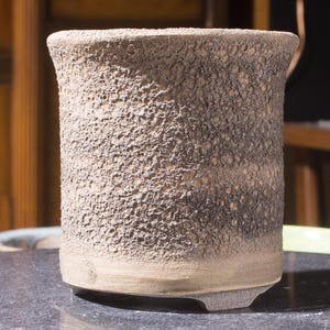May include: A cylindrical ceramic pot with a textured, speckled surface. The pot is mainly a light tan colour with darker brown and grey accents. The base is a lighter shade of tan, and the rim curves slightly outwards.