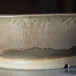 May include: A handmade ceramic bowl with a wide, shallow design. The bowl has a light brown exterior with a textured surface and a darker brown rim. The base is a lighter, off-white colour.