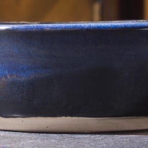 Może przedstawiać: Prostokątna ceramiczna miska z głębokim niebieskim szkliwem. Misa ma jasnobeżową podstawę z trzema małymi nóżkami. Niebieskie szkliwo przechodzi od jaśniejszego odcienia u góry do ciemniejszego odcienia u dołu.