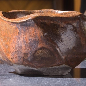 May include: A handcrafted, rustic ceramic bowl with an irregular shape. The bowl has a textured surface with brown, orange, and dark hues. The base is a lighter grey colour, adding contrast to the design. The bowl is approximately 15 cm wide.