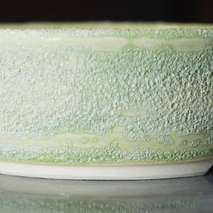 May include: A small, round ceramic bowl with a textured, pale green exterior. The bowl has a white base and a slightly uneven rim. The surface has a rough, speckled appearance, and is likely handmade.