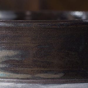 May include: A low, round ceramic bowl with a dark, mottled brown glaze. The bowl has a wide, shallow shape and a slightly textured surface. The rim is smooth, giving a rustic, handmade appearance. The bowl is suitable for serving.
