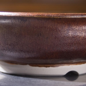 May include: A handmade ceramic bowl with a dark brown, glazed exterior and a white base. The bowl has a wide, shallow shape with a slightly flared rim. The interior is not visible.