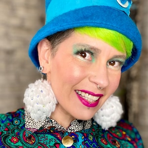 May include: A person with bright green hair is wearing a blue hat with a white button and a white pom-pom earring. They are wearing a colorful paisley patterned shirt.