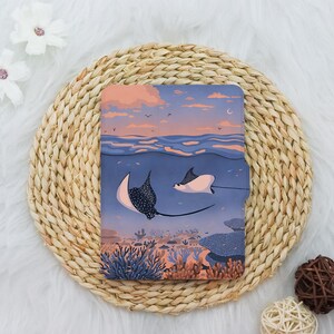 May include: A rectangular e-reader cover featuring a printed illustration of two spotted eagle rays swimming underwater. The scene depicts a sunset sky above the ocean surface, with coral reefs and sea plants visible on the ocean floor below.