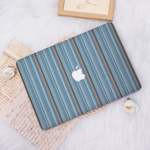 May include: A laptop with a blue, gray, and brown striped design. The Apple logo is visible on the laptop's surface. The laptop is placed on a woven mat, with a framed picture and a flower in the background.