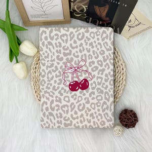 Könnte beinhalten: Eine Tablet-Hülle mit einem grauen Leopardenmuster. Die Hülle hat ein Design aus zwei roten Kirschen mit einer rosa Schleife. Die Hülle ist rechteckig und scheint aus einem strapazierfähigen Material zu sein.