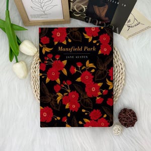 May include: A black hardcover book with a red floral design. The title "Mansfield Park" is printed in white on the front cover, along with the author's name, "Jane Austen".
