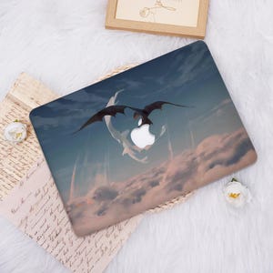 May include: Laptop case with a dragon design and a white Apple logo. The artwork shows a black dragon flying through a cloudy sky with blue, pink, and white colours. This case is a protective accessory for a laptop, designed to fit a specific model.
