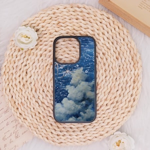 May include: A black phone case featuring a dreamy night sky design with clouds, stars, and a celestial aesthetic. The case is set against a woven, beige background with a white flower.