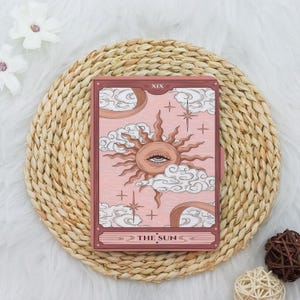 May include: A tarot card featuring a sun with an eye in the center, surrounded by clouds and stars, set against a pink background. The card is framed in a dark pink border and reads "THE SUN" at the bottom. The card is placed on a woven mat.