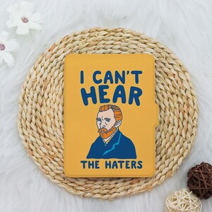 May include: Mustard yellow tablet case featuring a portrait of Vincent van Gogh and the text "I CAN'T HEAR THE HATERS" in blue. The case has a rectangular shape and is placed on a woven, circular placemat.