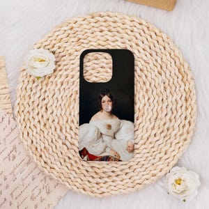 May include: Black phone case featuring a classical portrait of a woman blowing a bubble. The woman is wearing a white dress with puffed sleeves and a gold brooch. The case is on a woven, circular mat.
