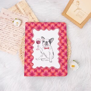 May include: A notebook with a pink and red retro pattern border. The center features a black and white French bulldog illustration holding a wine glass and wearing a red bow tie. The notebook is on a white surface.