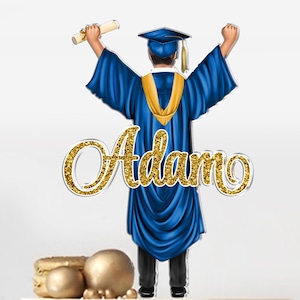 May include: A blue graduation cake topper with a gold cap and gown, holding a diploma. The topper is on a white cake with blue and black brushstrokes. The name "Adam" is written in gold glitter on the topper.