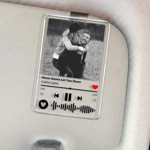 May include: Clear acrylic car accessory featuring a black and white photo of a couple. The design mimics a music player interface, with song title "Never Gonna Let You Down" and artist Colbie Caillat. Includes a red heart icon and a sound wave.