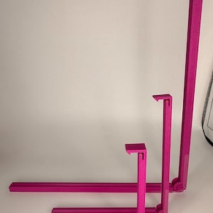 May include: Three magenta-colored, L-shaped shelf brackets of varying sizes are arranged vertically. The brackets are made of a solid material and have a textured surface. The brackets are designed to support shelves and provide a modern aesthetic.